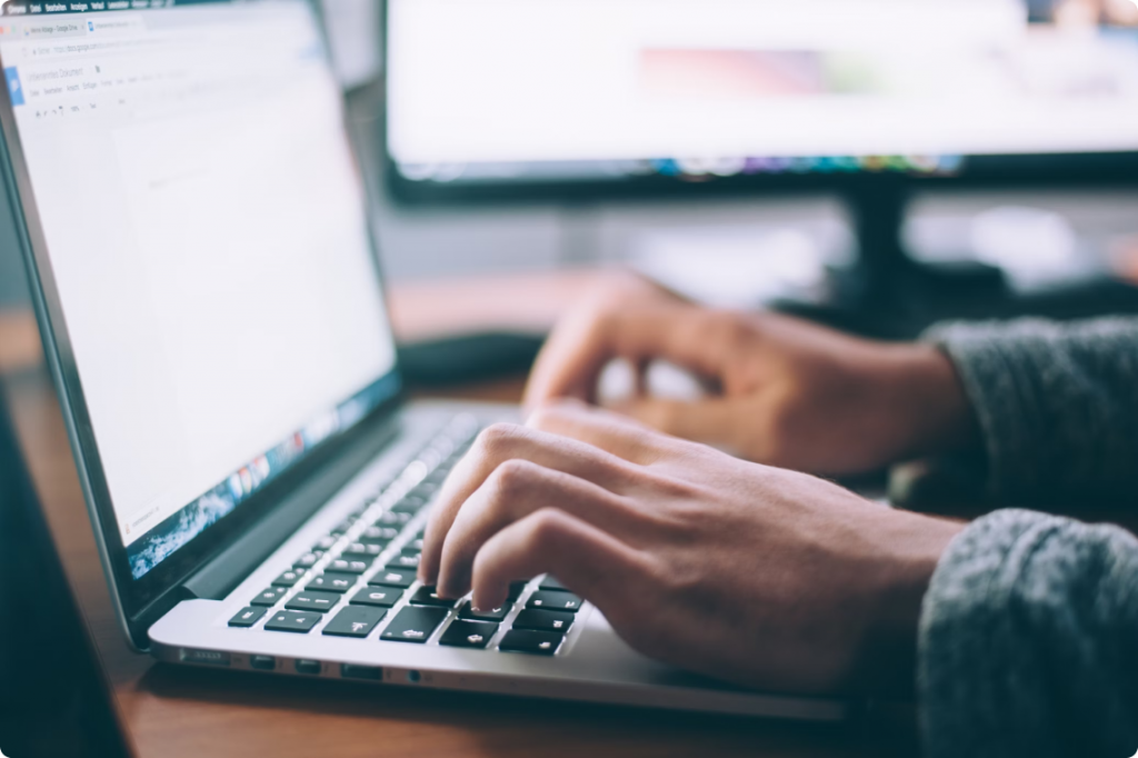 A pair of hands hovering over a laptop of a person sitting at desk with a desktop screen in the background 