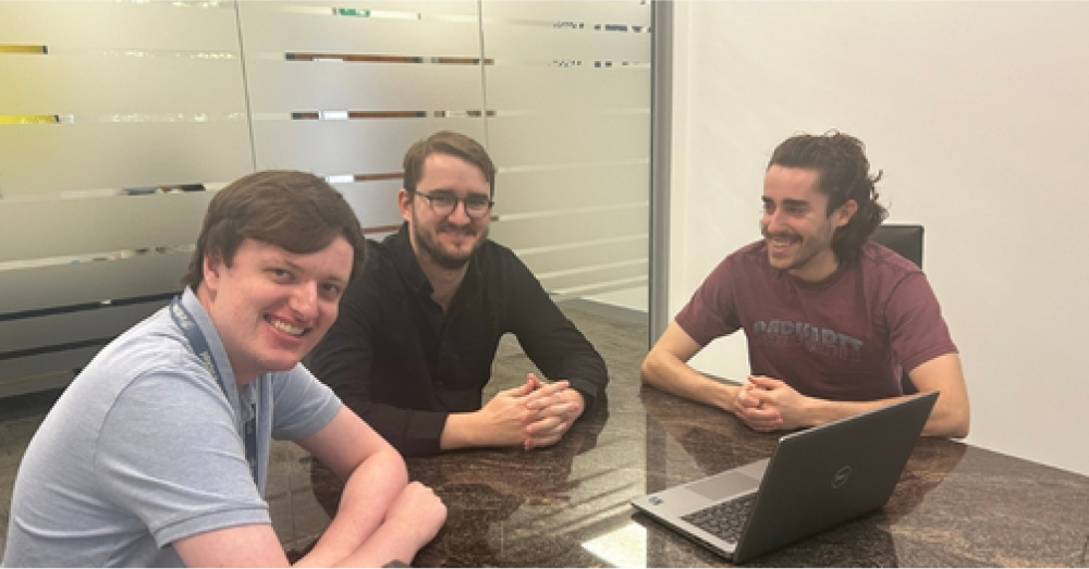 From left to right: Junior Developer Simon Horrocks, Data Scientist Sean Bayly and Junior Data Scientist Danny Glover sit at a table in a meeting room, looking happy and optimistic in front of a laptop.