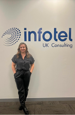 A white woman with blonde hair wearing a grey shirt and black trousers standing in front of a white wall with Infotel UK Consulting’s logo on it.